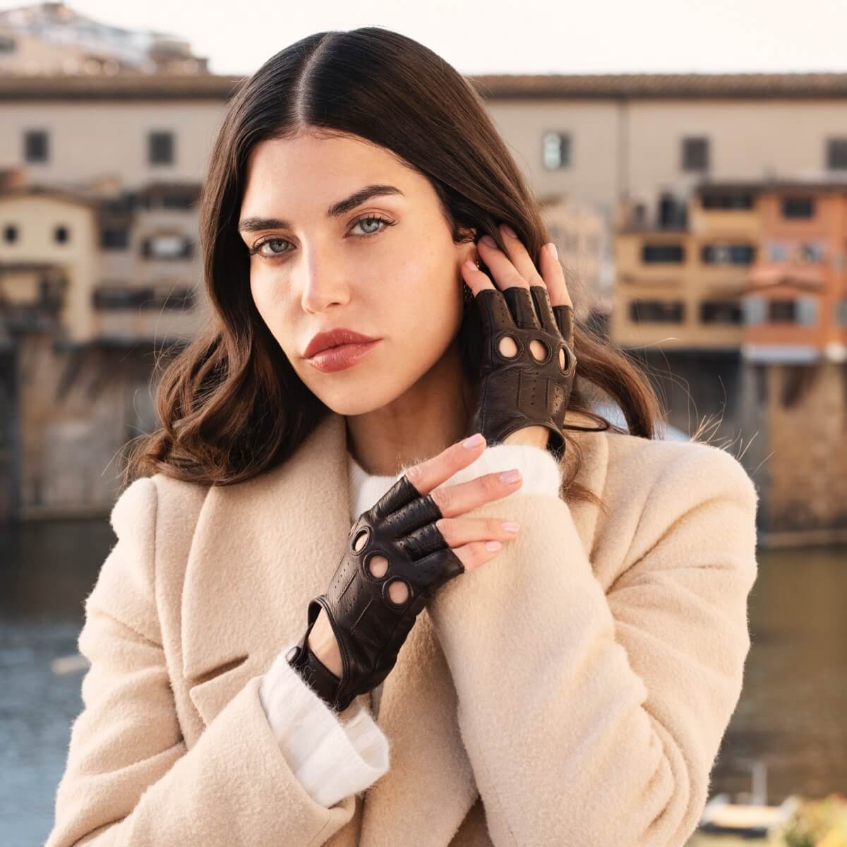 Woman wearing a beige coat and black fingerless gloves with a blurred cityscape background
