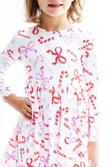 A child wearing a white long-sleeve dress with an allover holiday print of red and pink candy canes and bows, shown against a plain white background.