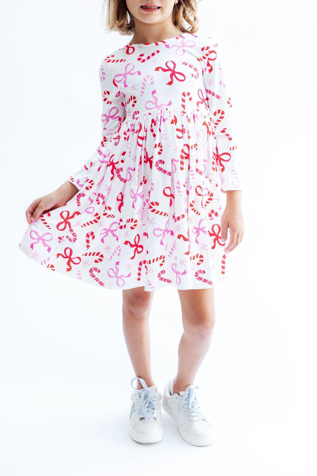 A young girl wearing a white long-sleeve dress with a red and pink candy cane and bow pattern, paired with white sneakers, standing against a plain white background.