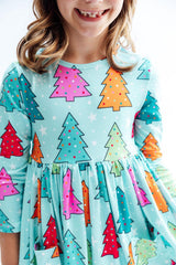 A close-up of a child wearing a light aqua long-sleeve dress with a colorful Christmas tree print in pink, orange, red, and green, set against a plain white background.