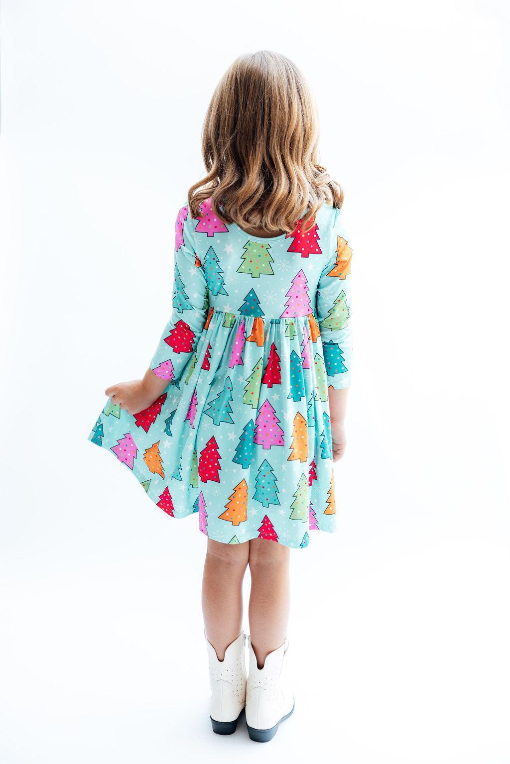 A young girl viewed from behind wearing a light blue-green dress with a colorful Christmas tree print, paired with white cowboy boots, against a plain white background.