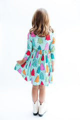 A young girl viewed from behind wearing a light blue-green dress with a colorful Christmas tree print, paired with white cowboy boots, against a plain white background.