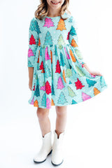 A young girl wearing a light blue tiered dress with a colorful Christmas tree print, paired with white cowboy boots, standing against a white background.