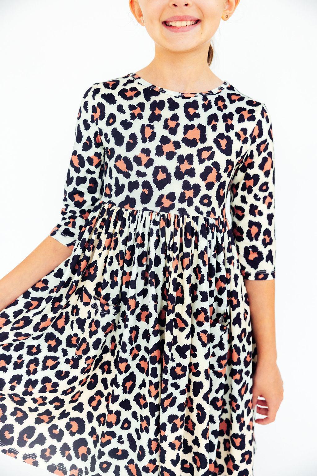 A child wearing a white three-quarter sleeve dress with a black and dusty rose leopard print, shown against a plain white background.