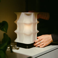 Person holding a textured white lamp on a surface with a plant in the background