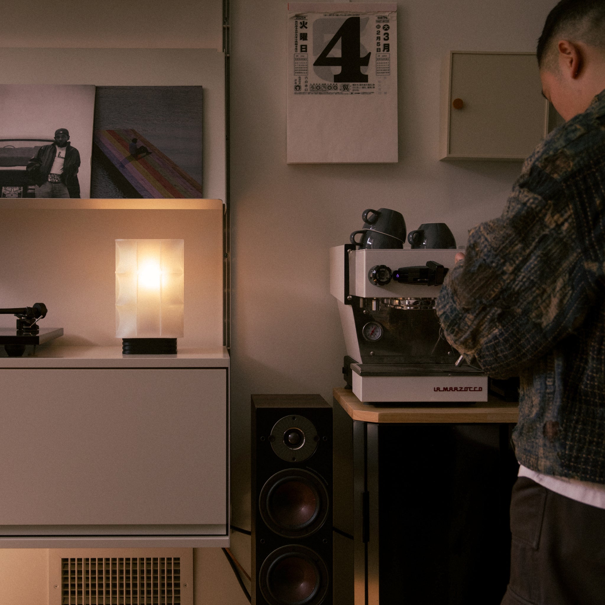 Person using a coffee machine in a room with speakers and a lamp.