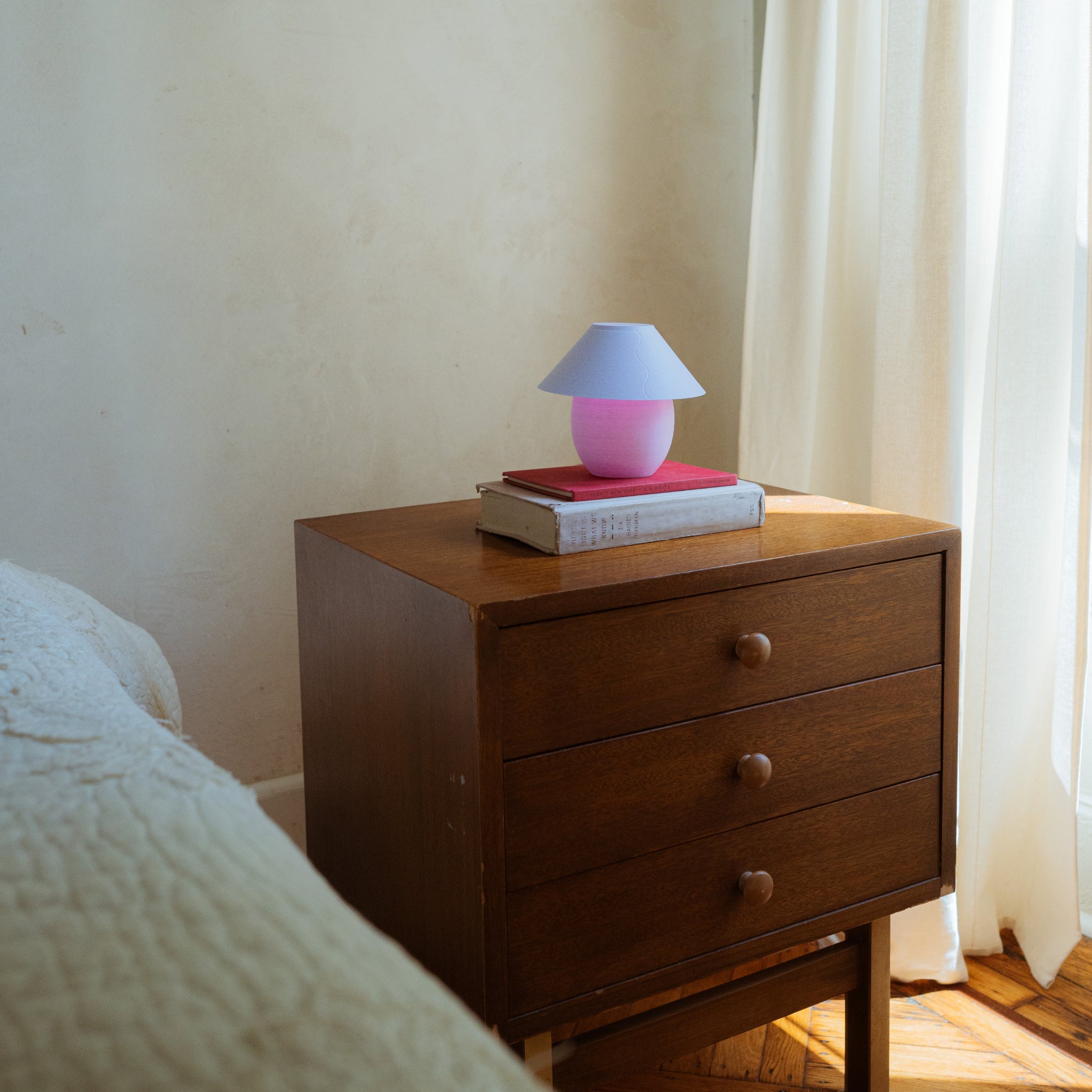 Wooden nightstand with a pink lamp and books next to a bed with white bedding.