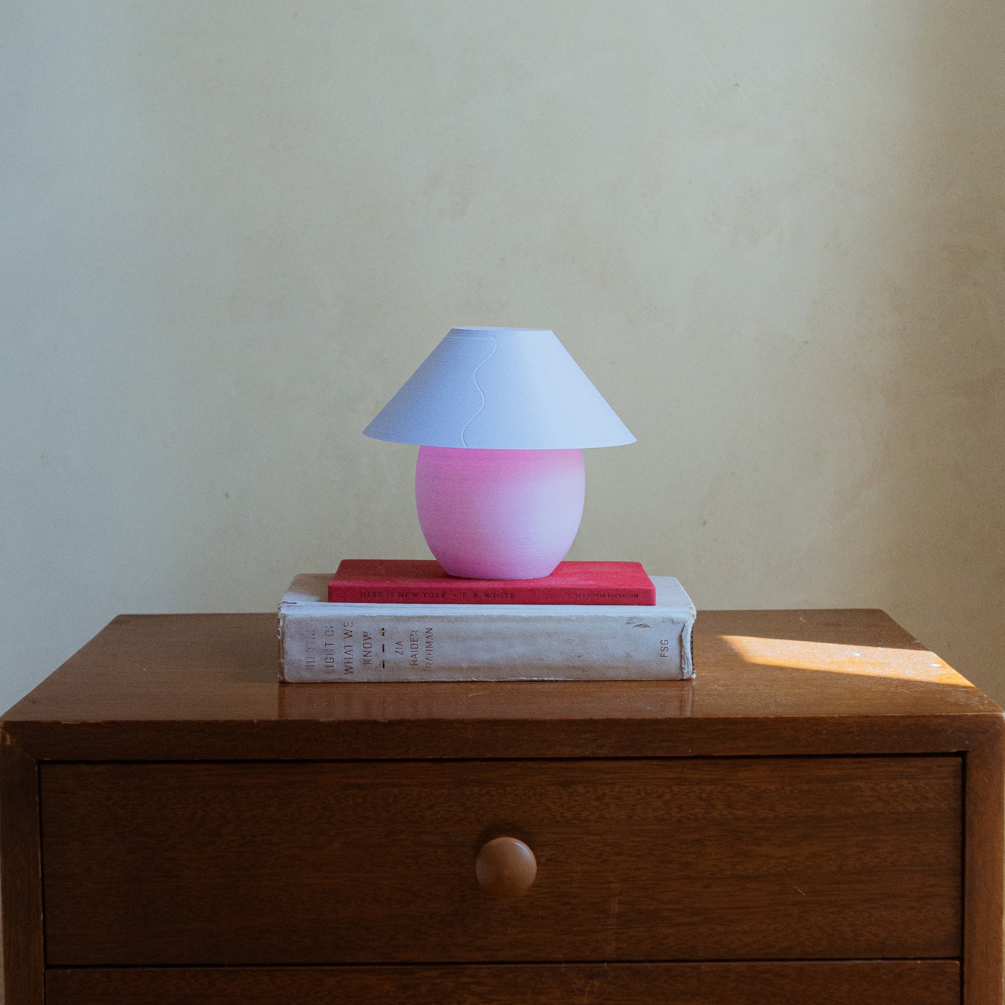 Pink lamp with a white shade on top of books on a wooden surface.