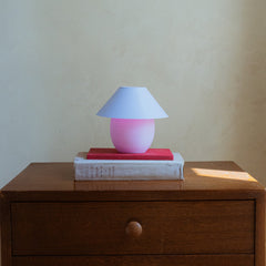 Pink lamp with a white shade on top of books on a wooden surface.