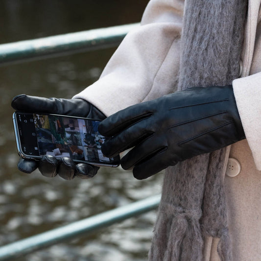 Black leather gloves worn by a person holding a smartphone, dressed in a cream coat and gray scarf with an outdoor railing in the background.