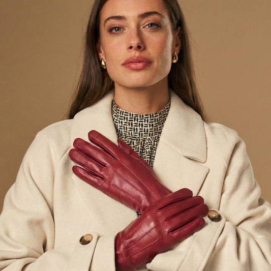 A woman wearing burgundy leather gloves over a cream-colored coat and a black-and-white geometric patterned top, posed against a solid beige background.