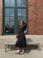 Woman in a black dress standing on a bench against a brick wall with large windows.