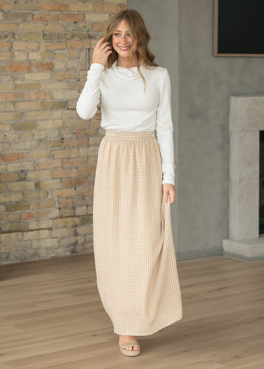 Woman wearing a white top and beige checkered skirt standing in a room with a brick wall and fireplace.