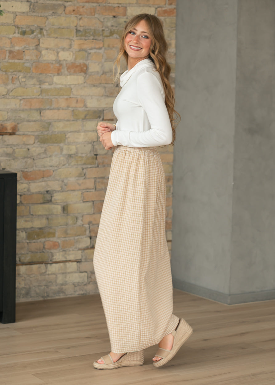 Woman wearing a white blouse and beige checkered pants standing against a brick wall.