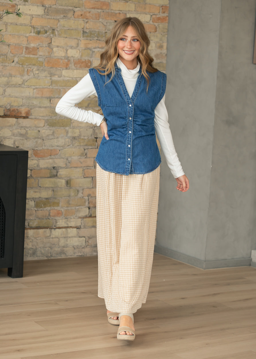 Woman wearing a blue vest over a white shirt with beige pants in a room with a brick wall.