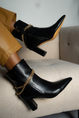 Black ankle boots with gold chains worn by a person sitting on a beige couch.