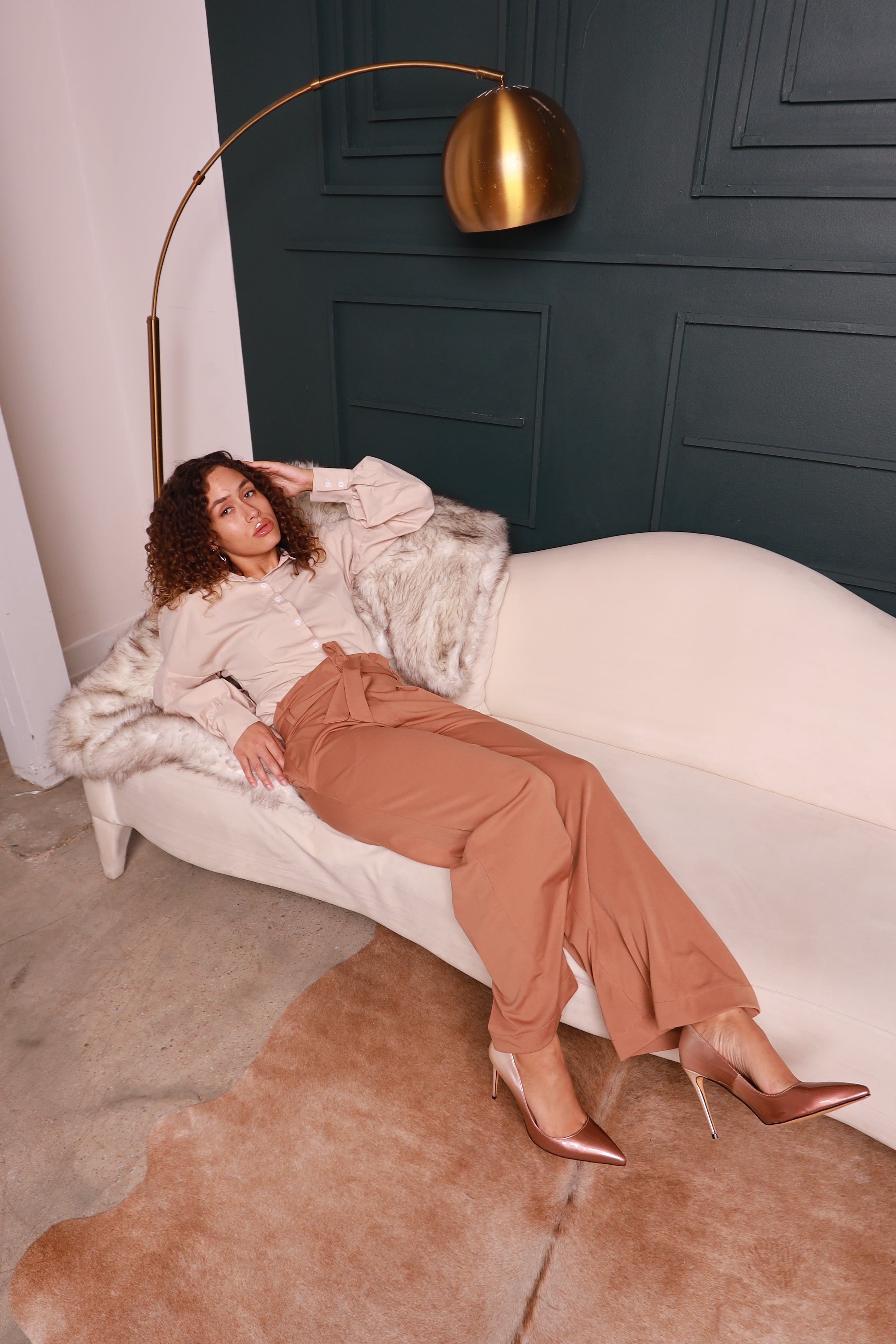 Woman in a stylish outfit sitting on a white chair in a room with dark walls and a gold floor lamp.