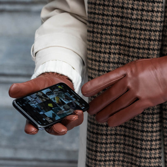 A close-up of a person wearing brown leather gloves, a cream jacket, and a brown and beige houndstooth coat, holding a smartphone.