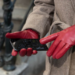 A close-up of a person wearing bright red leather gloves and a beige wool coat, holding a smartphone with the gloves.