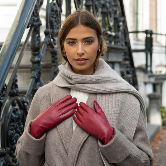 A woman wearing a beige wool coat, a light gray knit scarf, and bright red leather gloves, standing in front of an ornate black metal staircase.