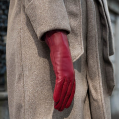 A close-up of a person wearing a long, structured taupe wool coat and a vibrant red leather glove on one hand.