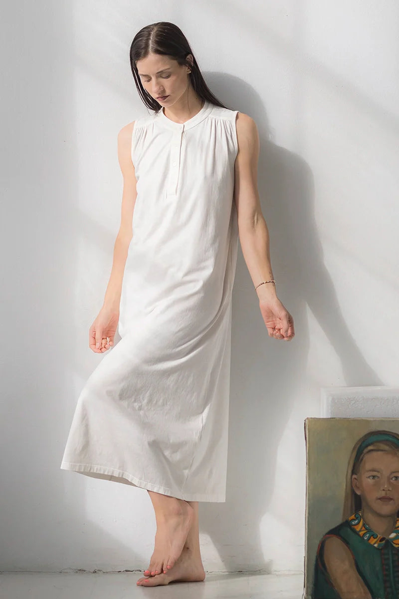 Woman wearing a white sleeveless dress standing against a plain white wall.