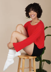 Woman in a red sweater and black pants sitting on a wooden stool against a beige background