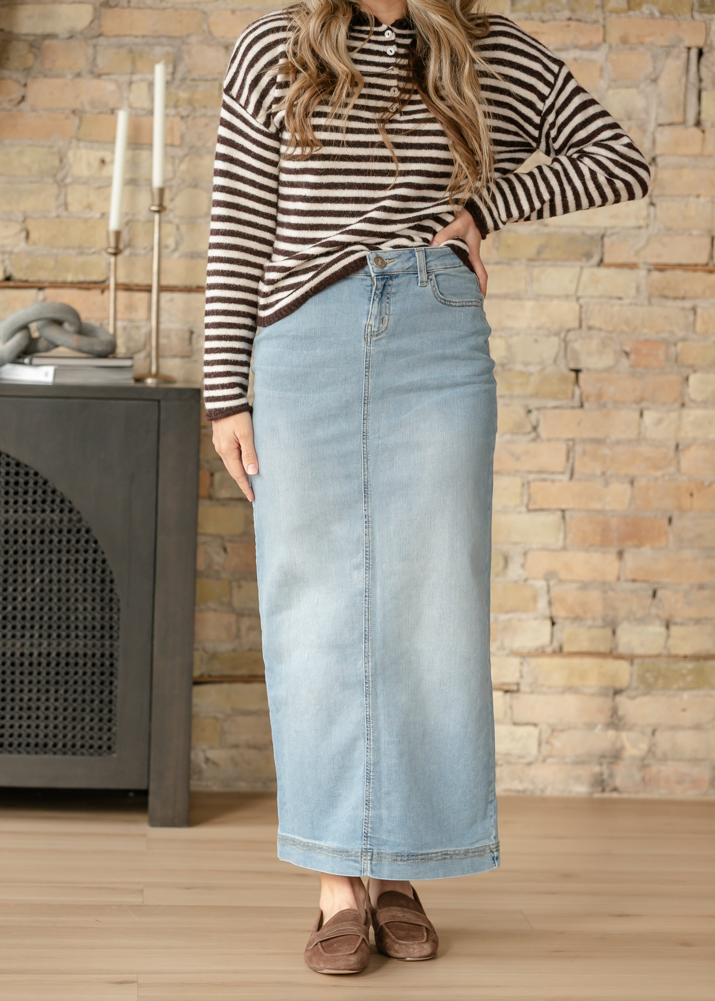 Person wearing a striped long-sleeve top and denim skirt standing in front of a stone wall.