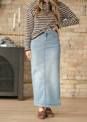 Person wearing a striped long-sleeve top and denim skirt standing in front of a stone wall.