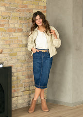 Woman wearing a white blouse, beige jacket, blue denim skirt, and brown boots against a brick wall.