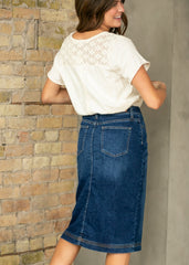 Woman wearing a white lace top and blue denim skirt against a stone wall.