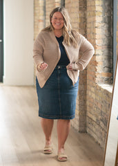 Woman wearing a beige cardigan and blue denim skirt standing in a room with brick walls.