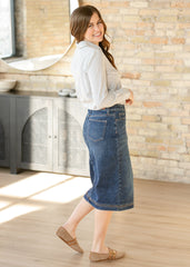 Woman wearing a white blouse and blue denim skirt standing in a room with a stone wall.
