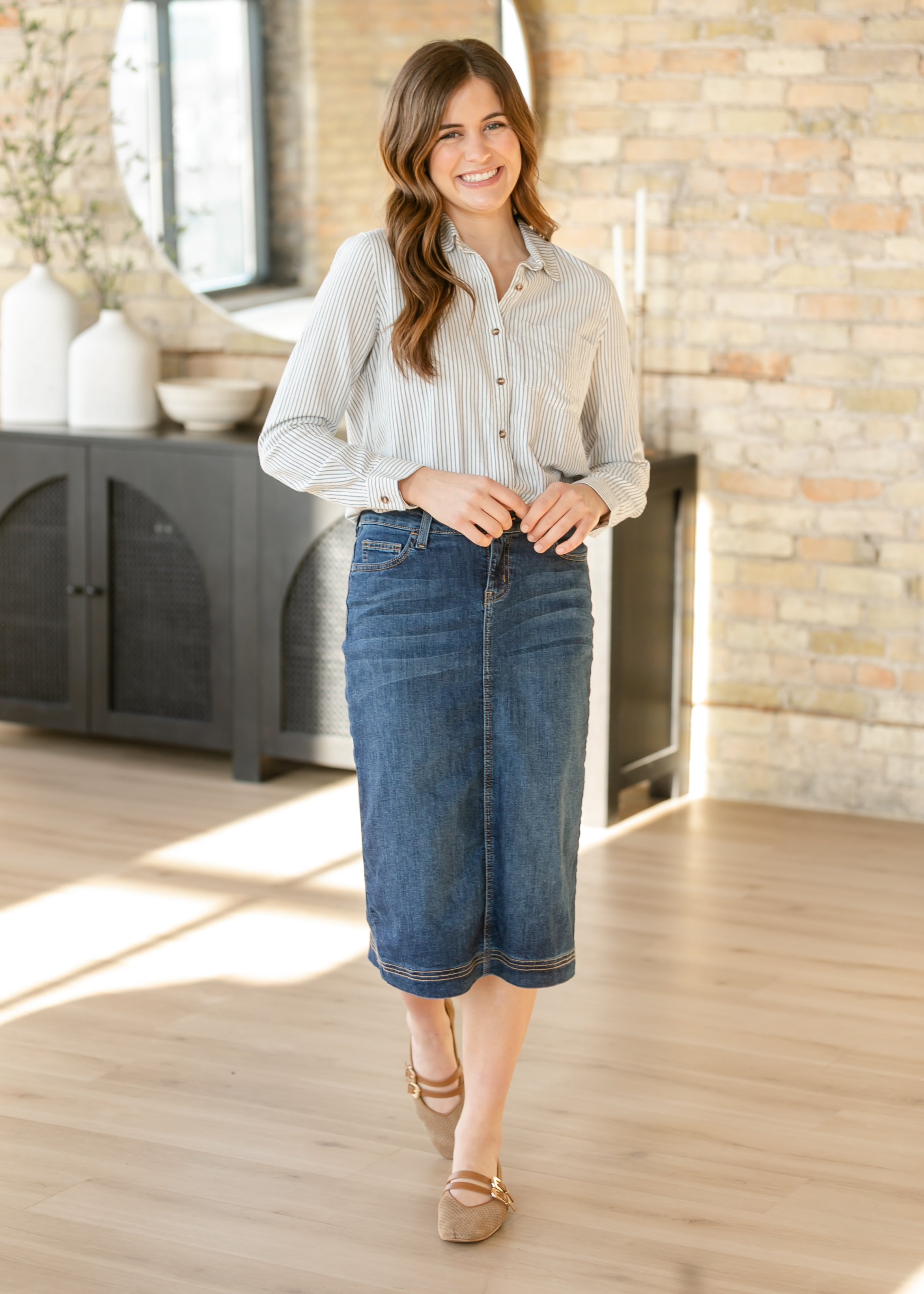 Woman wearing a light-colored shirt and blue denim skirt in a room with wooden flooring and a stone wall.