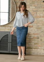 Woman wearing a light gray blouse and blue denim skirt standing against a brick wall.
