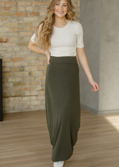 Woman wearing a white top and olive green skirt standing in a room with a brick wall.