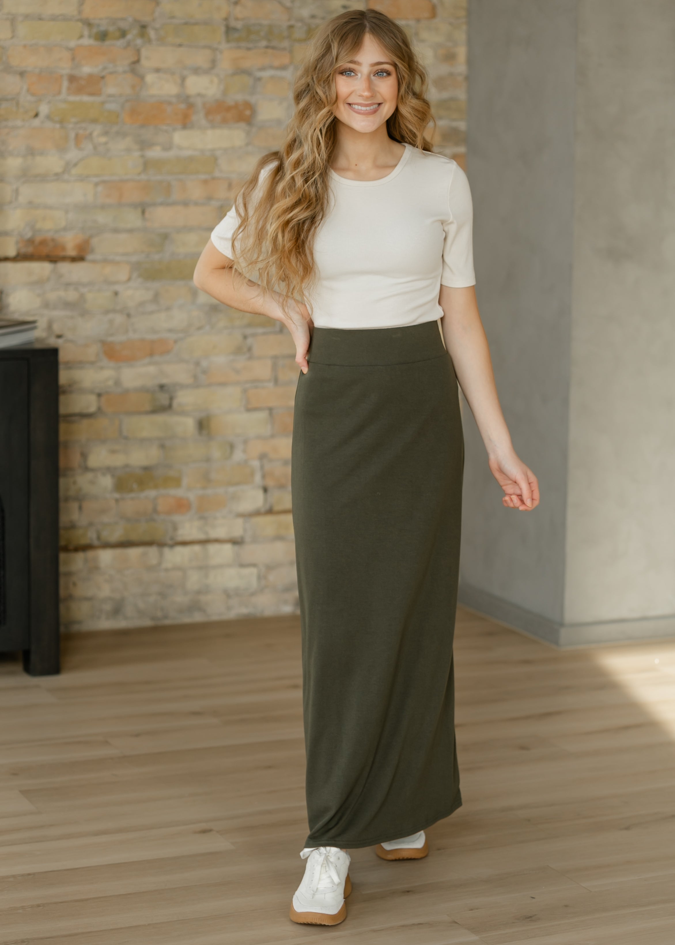 Woman wearing a white top and long green skirt standing in a room with a stone wall.