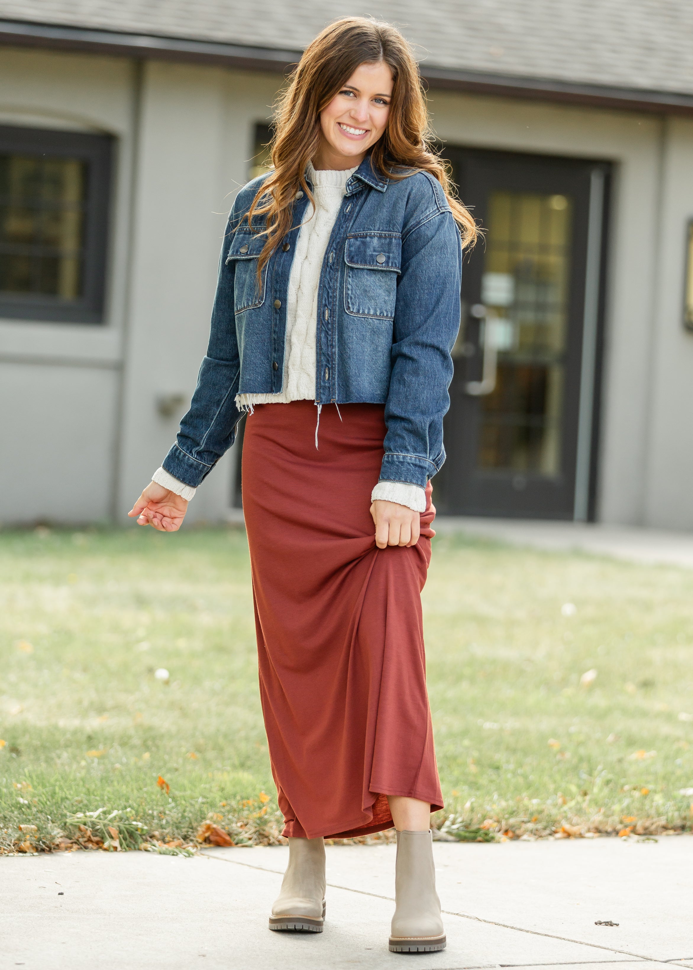 Woman wearing a blue denim jacket, white sweater, red skirt, and beige boots standing outside a house.