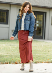 Woman wearing a blue denim jacket, white sweater, red skirt, and beige boots standing outside a house.