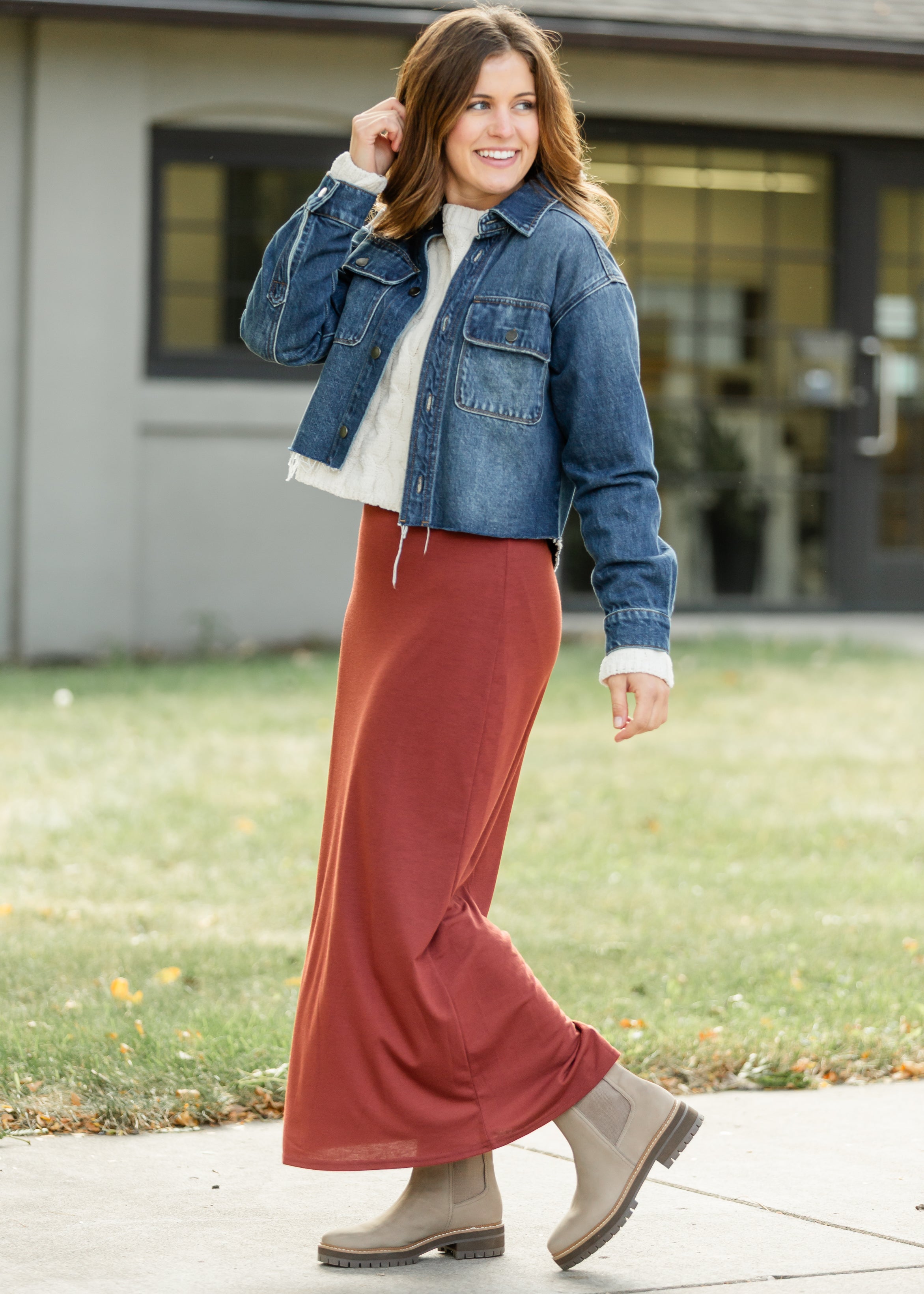 Woman wearing a denim jacket, white shirt, rust-colored pants, and brown boots outdoors.