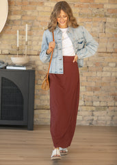 Woman in a denim jacket and brown skirt standing in a room with a stone wall.