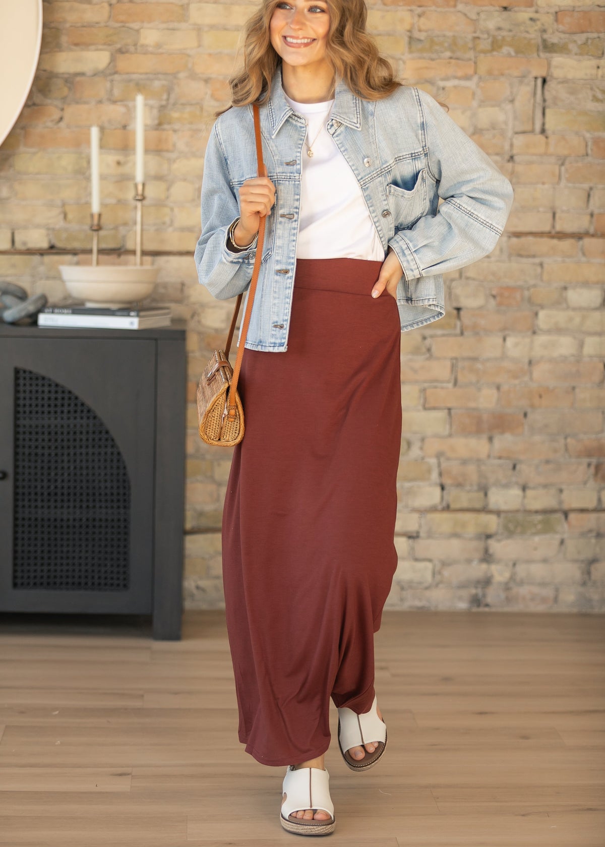 A woman models a light-wash blue denim jacket and a long, flowing rust-brown maxi skirt, styled with a white t-shirt, a small brown crossbody bag, and white slide sandals. She stands in a room with a rustic brick wall background.