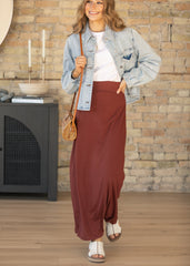 A woman models a light-wash blue denim jacket and a long, flowing rust-brown maxi skirt, styled with a white t-shirt, a small brown crossbody bag, and white slide sandals. She stands in a room with a rustic brick wall background.