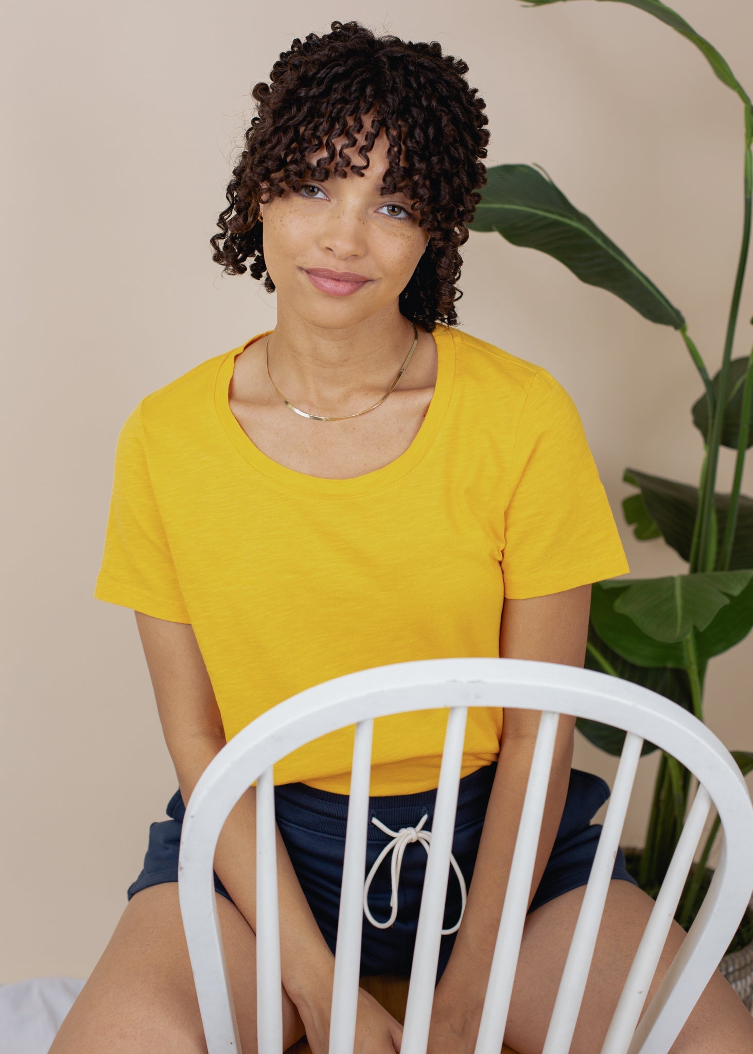 Woman in a yellow shirt sitting on a white chair with a plant in the background