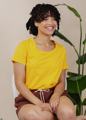 Woman wearing a yellow shirt and brown shorts sitting in front of a plant.