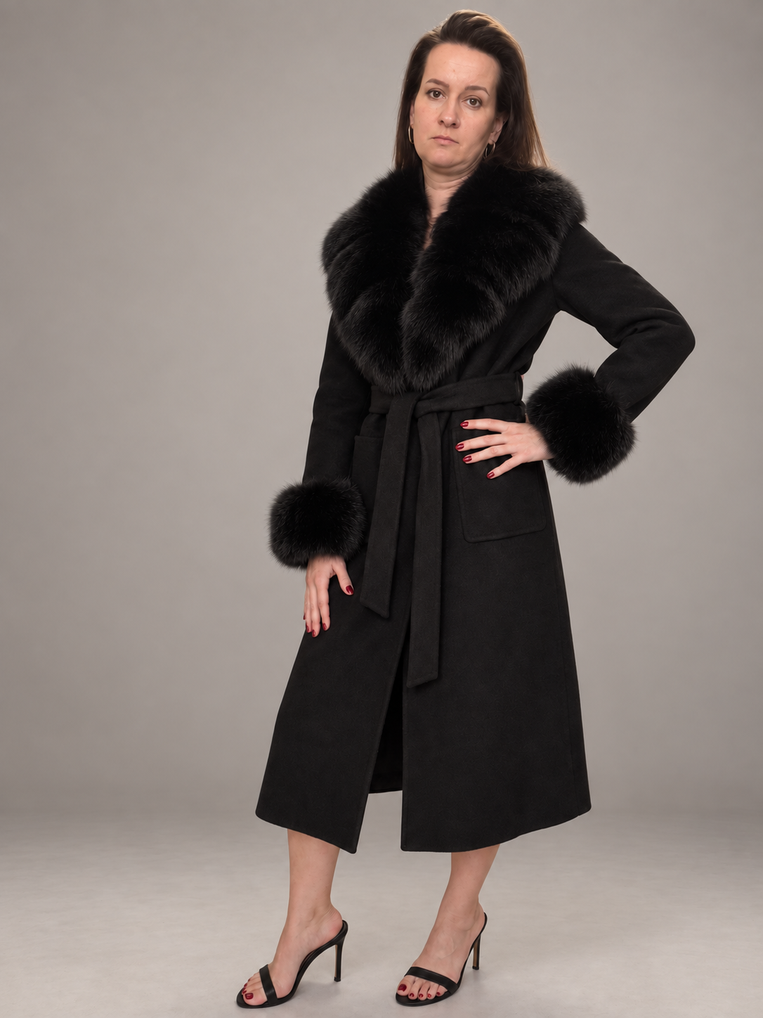A woman modeling a black mid-length coat with a large black fur collar, matching belt, and fur-trimmed cuffs, paired with black heeled sandals against a plain gray studio background.