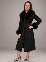 A woman modeling a black mid-length coat with a large black fur collar, matching belt, and fur-trimmed cuffs, paired with black heeled sandals against a plain gray studio background.
