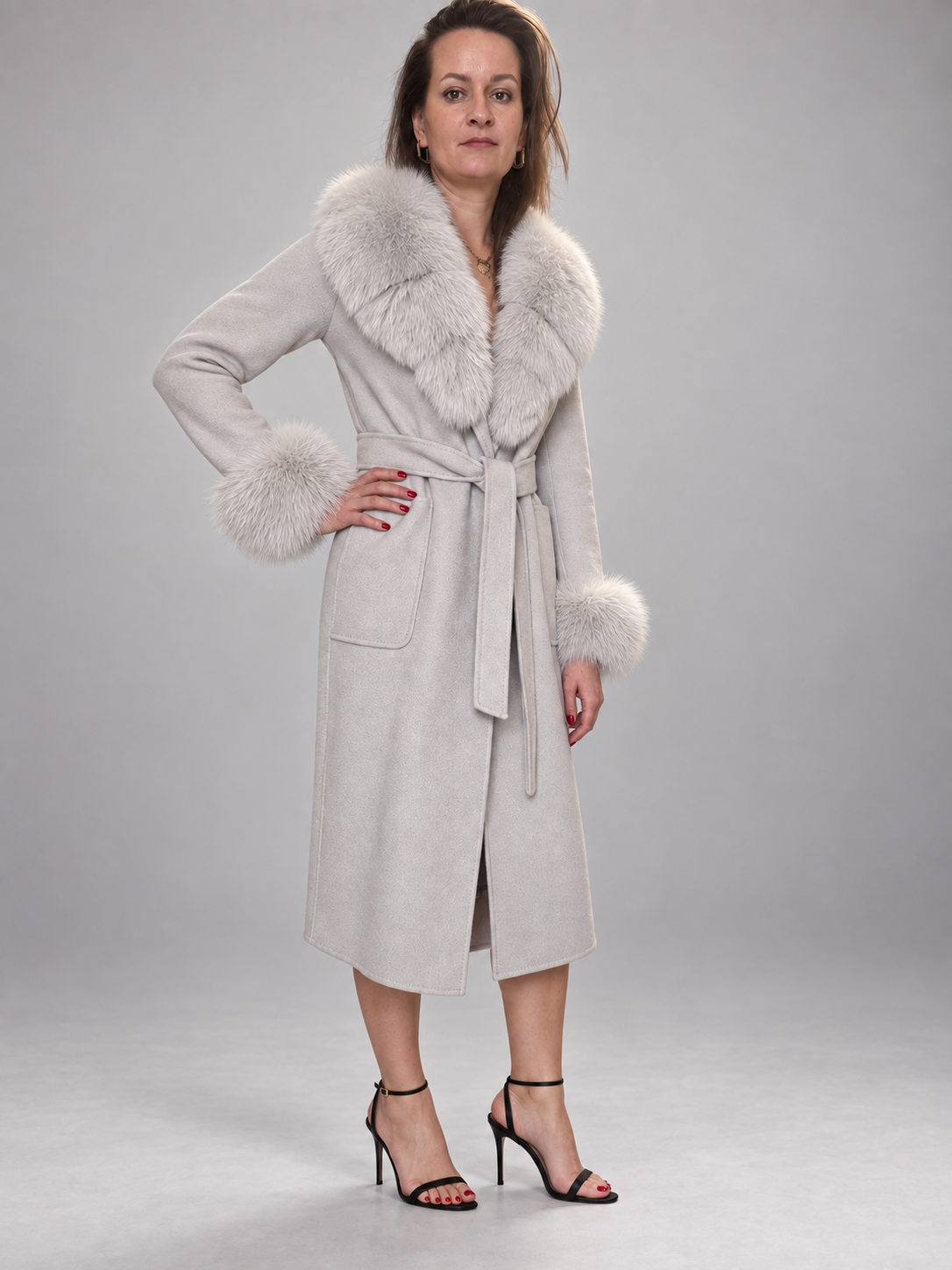A woman models a light beige faux-suede wrap coat with a large white fur collar and matching fur-trimmed cuffs, paired with black high-heeled sandals against a neutral gray studio background.