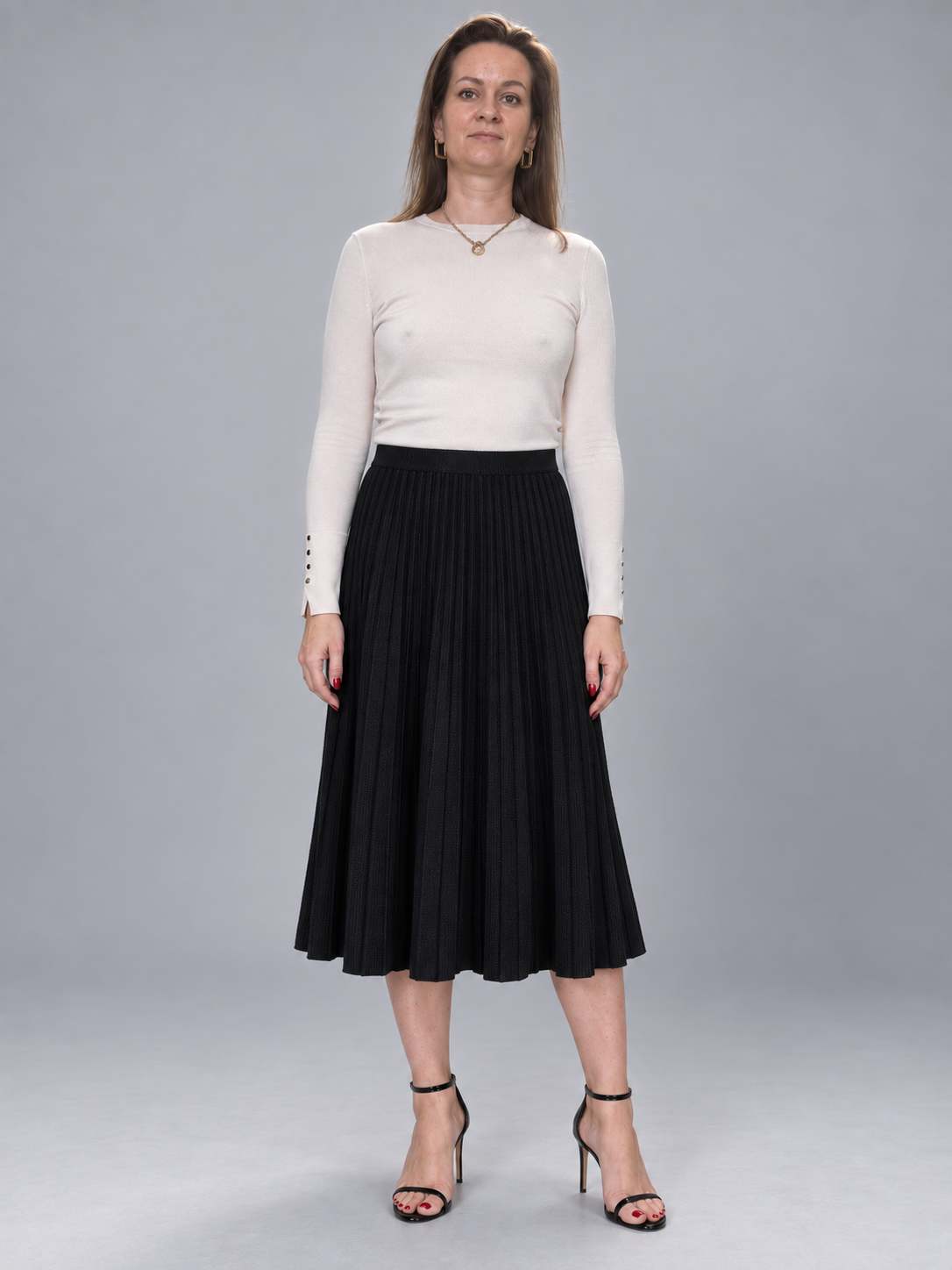 A woman modeling a cream long-sleeve knit top and a black pleated midi skirt, paired with black strappy high-heeled sandals, against a solid gray studio background.
