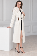 A woman models a long cream-colored coat with a plush fur collar, a matching belt with a brown buckle, and a black dress underneath, standing in a modern room with a white block pedestal.